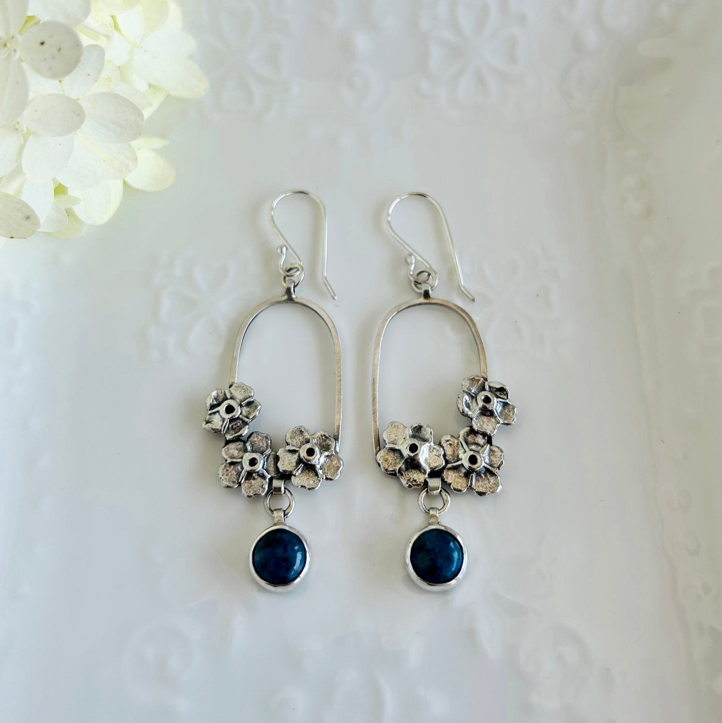 Silver earrings with floral designs and blue stones on a white textured background