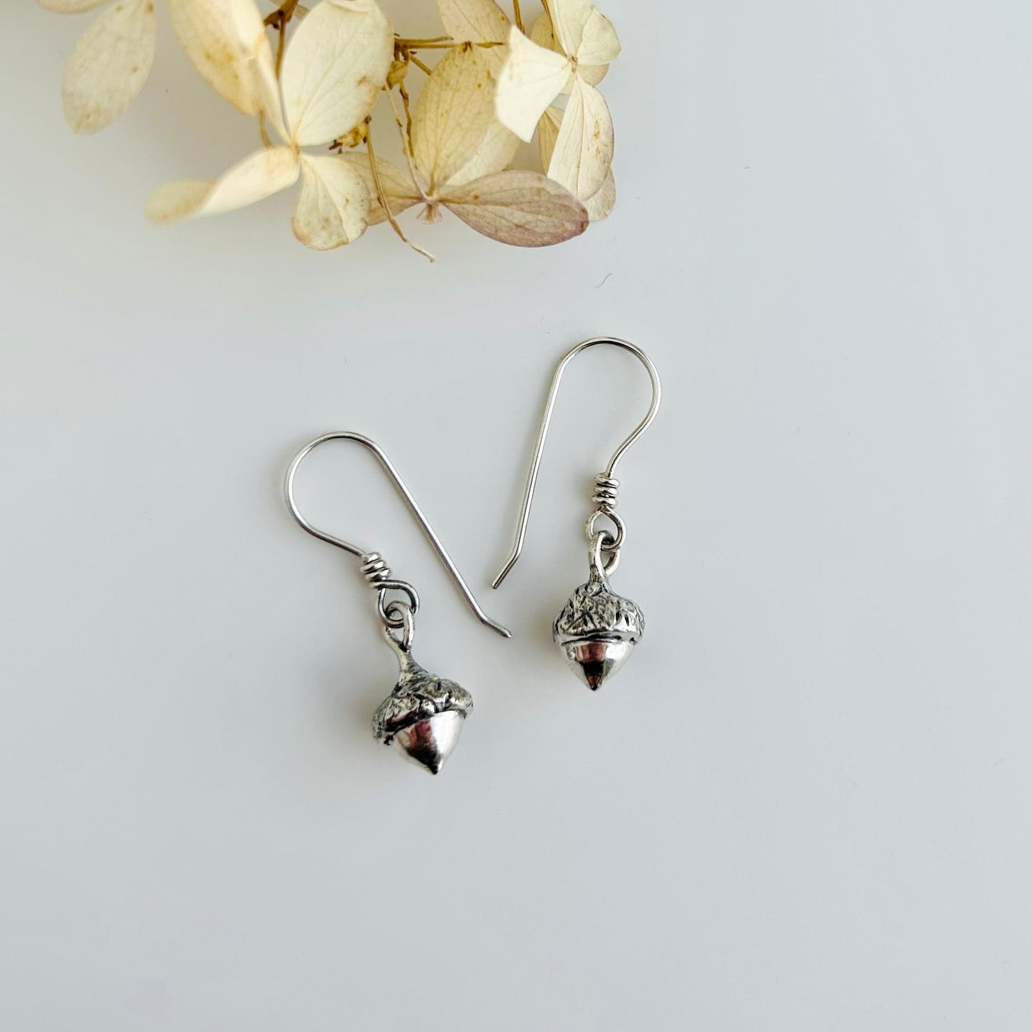 Silver acorn-shaped earrings on a light gray background with dried flowers.
