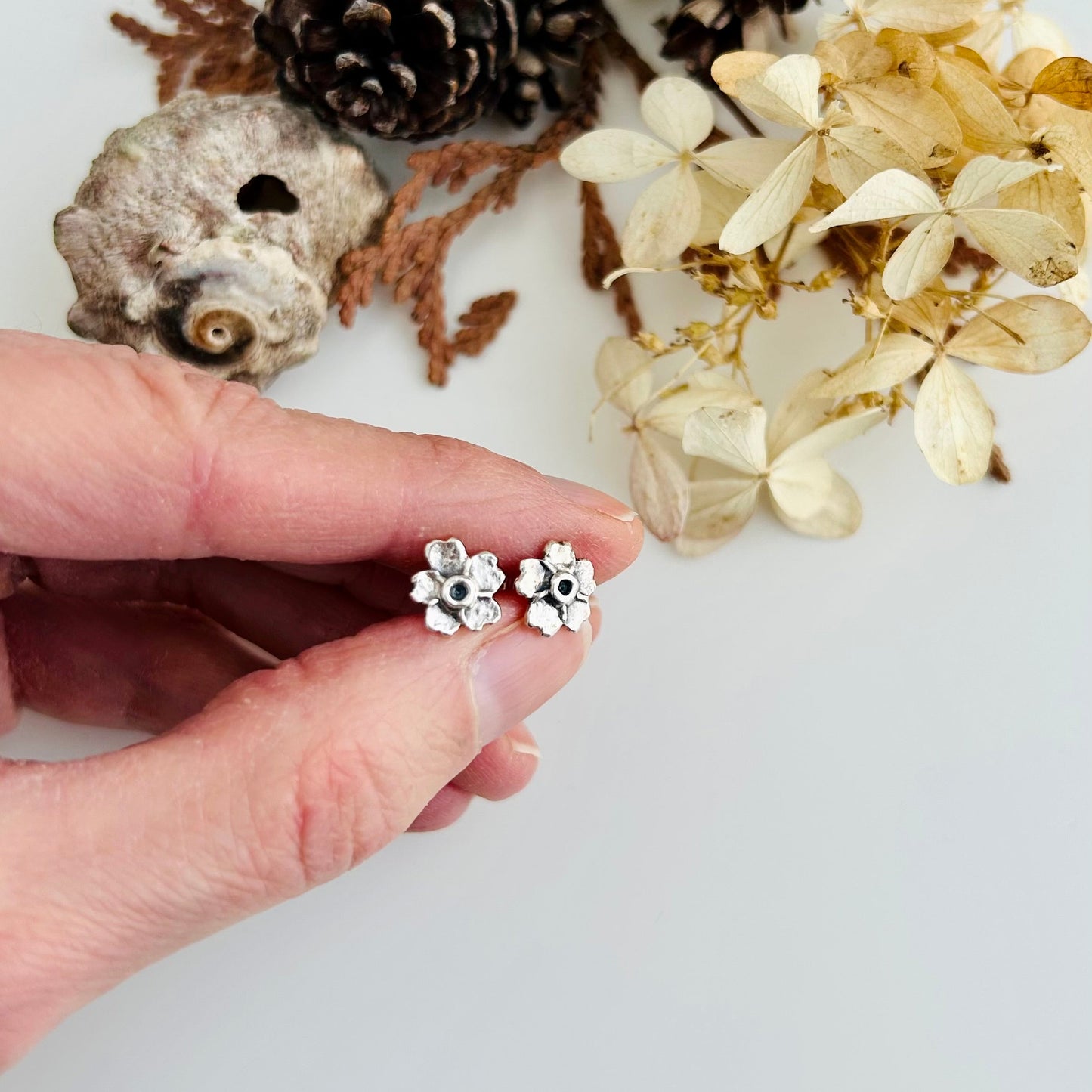 Hand holding a pair of silver flower-shaped earrings with a natural background of pinecones and dried flowers.