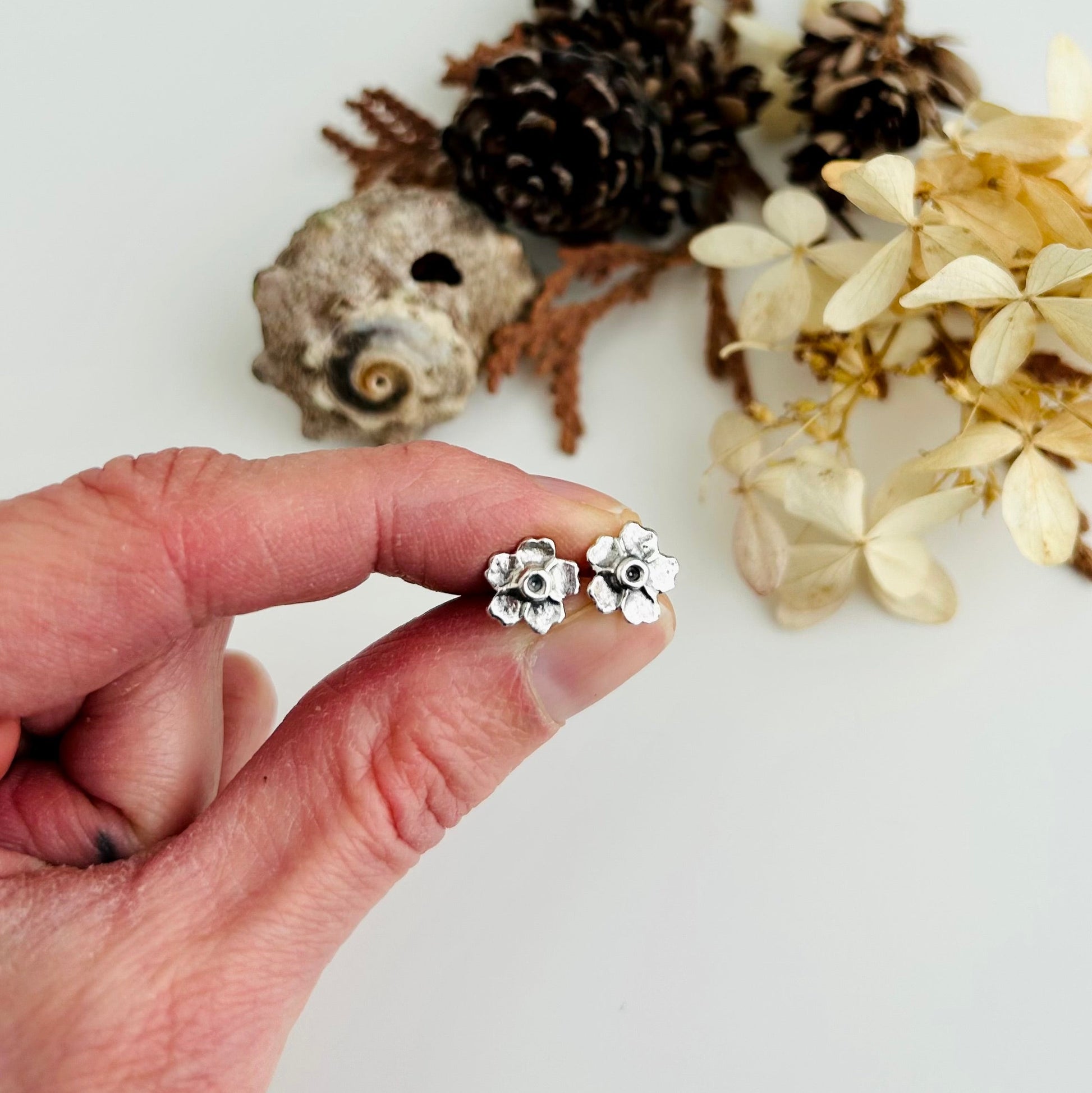 Hand holding a pair of silver floral earrings with natural elements in the background.